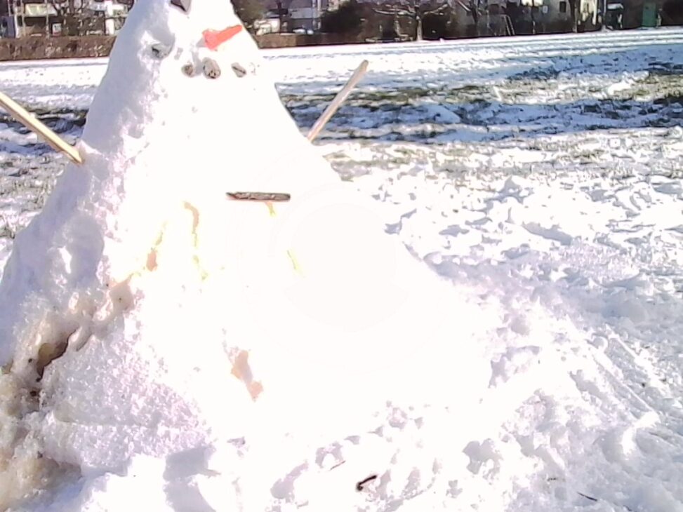 Small snowman built on a snow-covered field, with a carrot nose, pebble eyes, and stick arms, photographed in harsh sunlight that overexposes parts of the snow; buildings and trees are faintly visible in the background beyond the field.