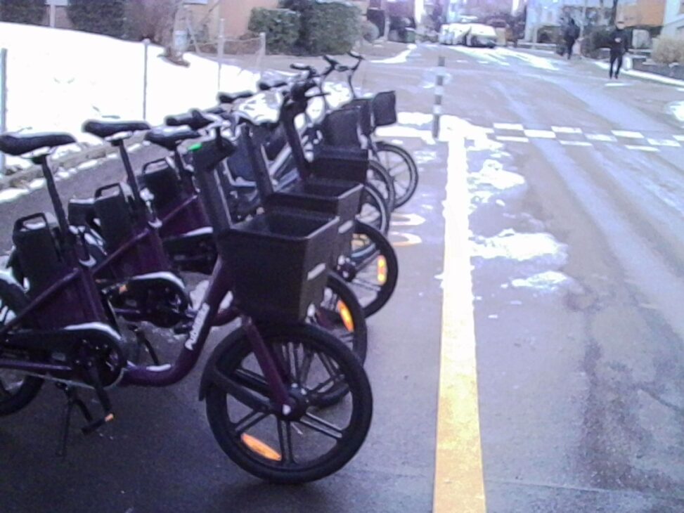 Row of parked rental bicycles with front baskets lined up along a wet residential street, patches of snow and ice on the ground, a yellow road marking running alongside the bikes, and a pedestrian visible in the distance under overcast winter light.
