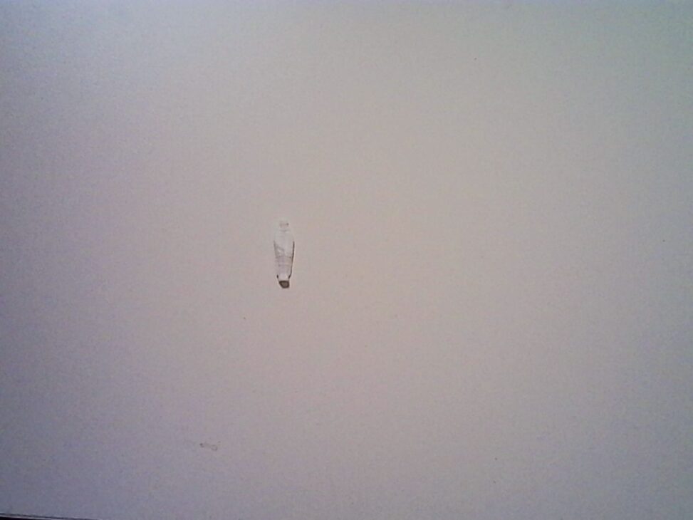 Small plastic figurine from a King’s cake resting on a plain, light-colored surface, centered in the frame with soft, uneven lighting and visible digital noise.