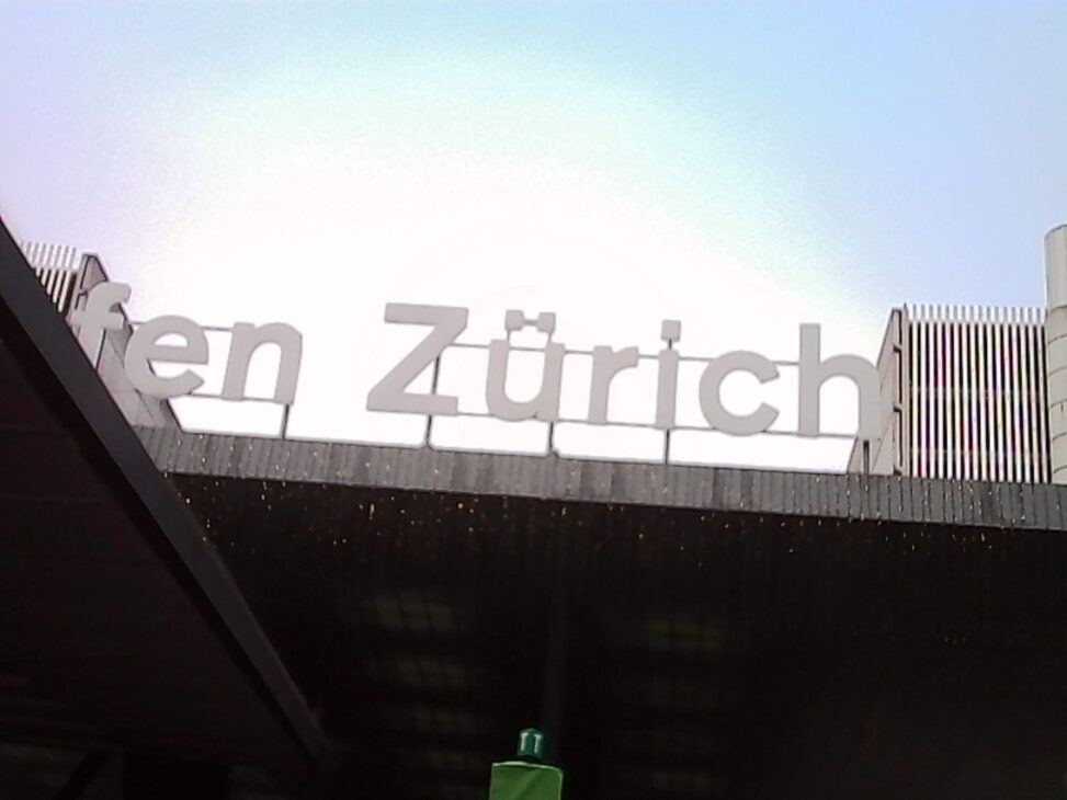 Large white “Zürich” lettering mounted on the edge of a concrete structure at the airport, photographed from below against a pale, overexposed sky, with architectural elements and vertical slats visible on either side.