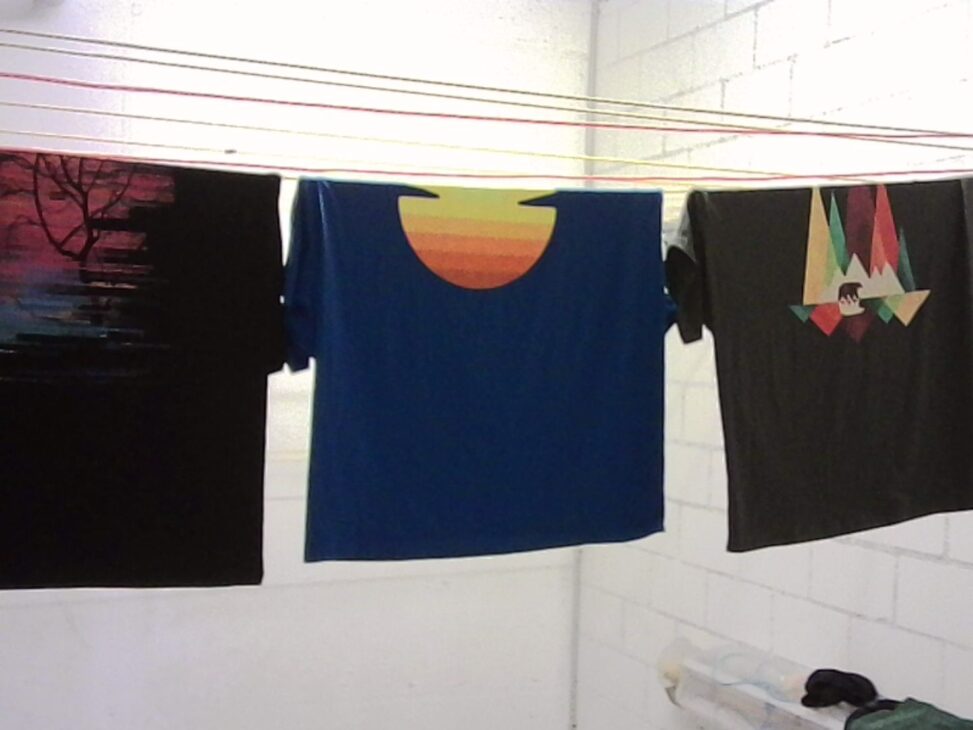 Three graphic t-shirts hanging to dry indoors on parallel clotheslines, photographed against white tiled walls, with colorful printed designs visible on the shirts and soft, uneven indoor lighting.