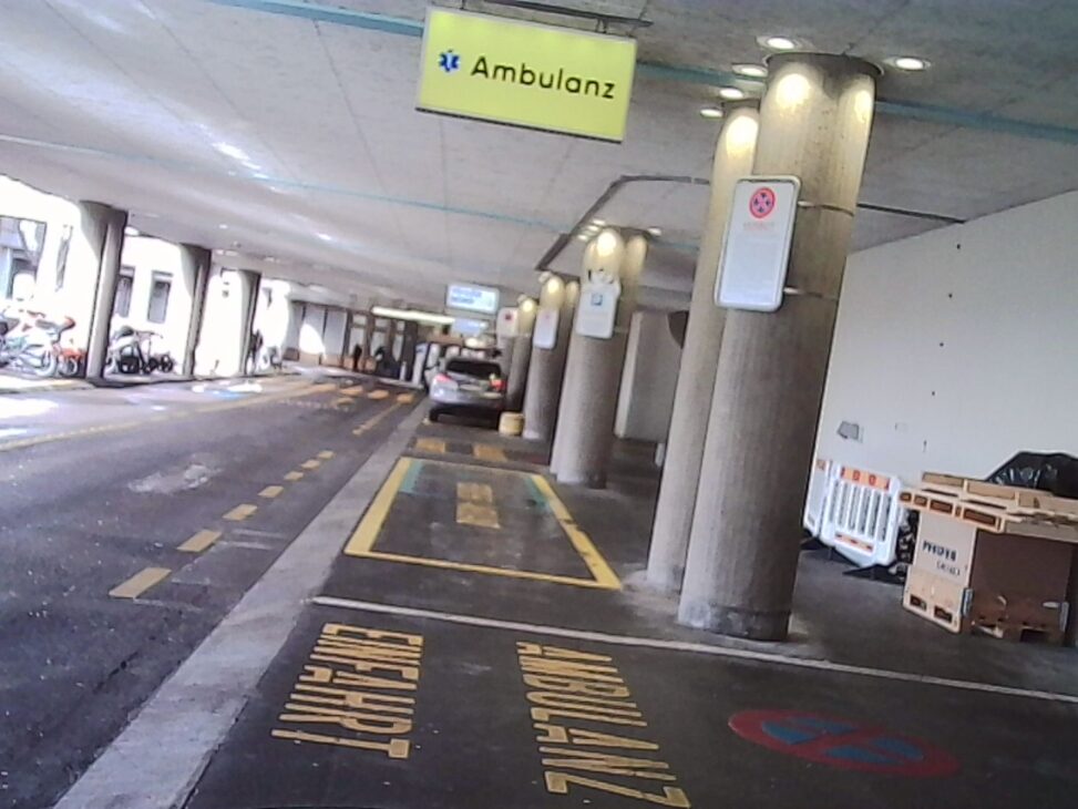 Covered hospital driveway with concrete pillars and marked parking lanes, an “Ambulanz” sign hanging overhead, a parked car in the distance, and yellow road markings on wet pavement under artificial lighting.