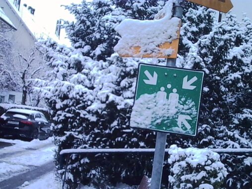 Snow-covered residential street with evergreen trees and hedges, a green emergency assembly point sign partially covered in snow mounted on a metal pole, a parked car to the left, and compacted snow and slush on the road under overcast winter light.
