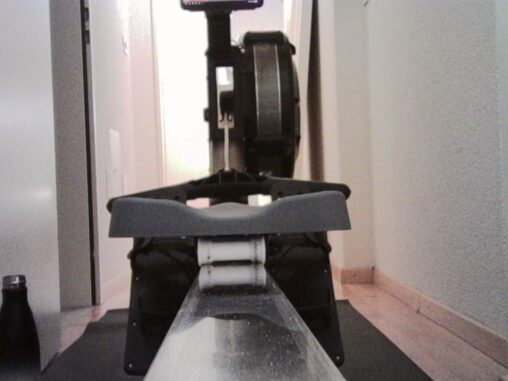 Close-up, front-facing view of an indoor rowing machine, with the seat, footrests, and rail aligned symmetrically, set in a narrow hallway or room with pale walls and soft, even lighting.