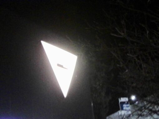 Night-time scene with a brightly illuminated triangular transport sign glowing against a dark sky, partially framed by bare tree branches, with a dimly lit tram stop and signage visible in the background.