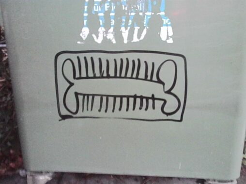 Close-up of a simple black line drawing on a pale surface, depicting a rectangular frame containing two bone-like shapes with short vertical strokes between them, photographed in diffuse daylight with slight surface texture visible.