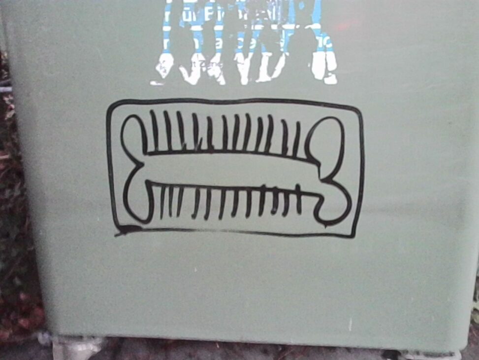 Close-up of a simple black line drawing on a pale surface, depicting a rectangular frame containing two bone-like shapes with short vertical strokes between them, photographed in diffuse daylight with slight surface texture visible.