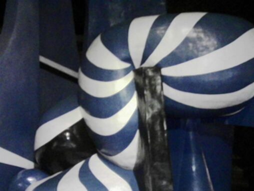 Close-up of a sculptural surface with alternating blue and white curved stripes, glossy and slightly reflective, photographed at night under artificial light, with dark surroundings fading into shadow.