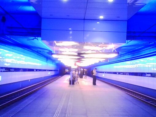 Underground streetcar platform in Zurich with a strong blue light installation along tiled walls, creating a tunnel-like perspective; a few people stand spaced out on the platform beneath geometric ceiling lights, with tracks running on both sides and the scene softly blurred by low-light conditions.