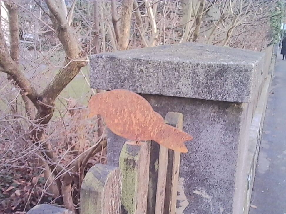 A small, rusted metal bird figurine mounted on a weathered stone and wooden structure beside a sidewalk, surrounded by bare winter branches.