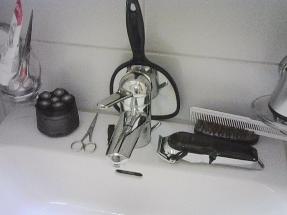 A bathroom sink with a chrome faucet surrounded by grooming tools, including scissors, hair clippers, brushes, and a razor, arranged neatly on a white surface.