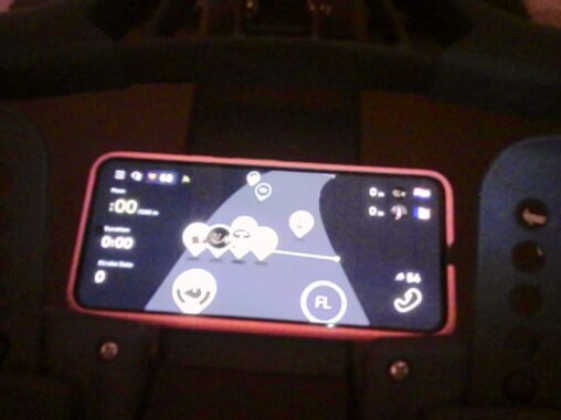 A smartphone mounted horizontally on a rowing machine displays a virtual rowing interface with multiple avatar markers on a map, representing other users rowing simultaneously. The phone rests against a dark padded surface in a dimly lit indoor setting.