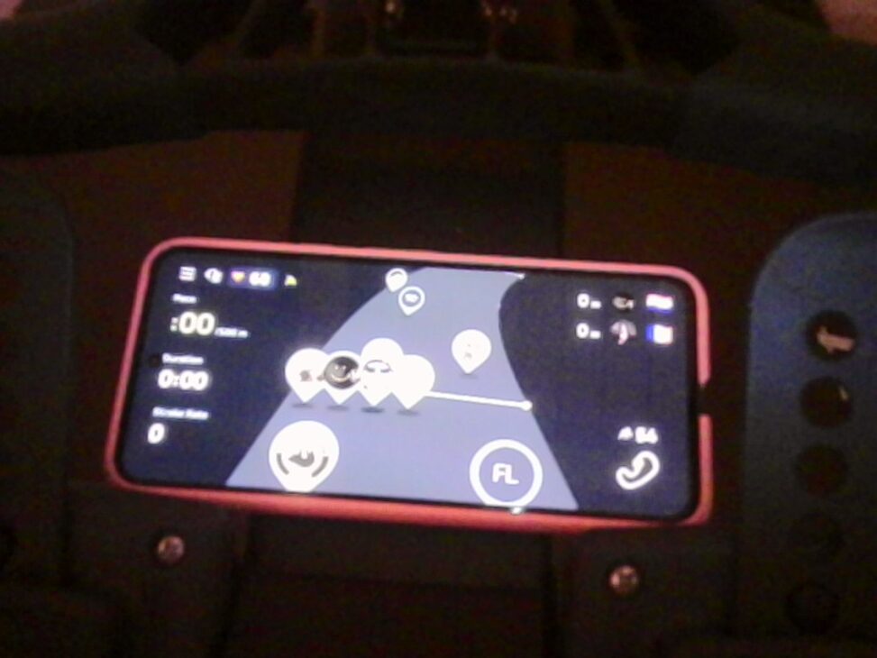 A smartphone mounted horizontally on a rowing machine displays a virtual rowing interface with multiple avatar markers on a map, representing other users rowing simultaneously. The phone rests against a dark padded surface in a dimly lit indoor setting.