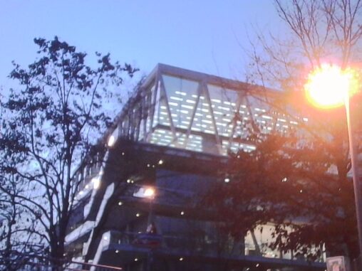 Modern multi-level glass-and-steel building at dusk in Zurich, illuminated from within, with diagonal supports visible through light fog and a warm streetlight glowing in the foreground.