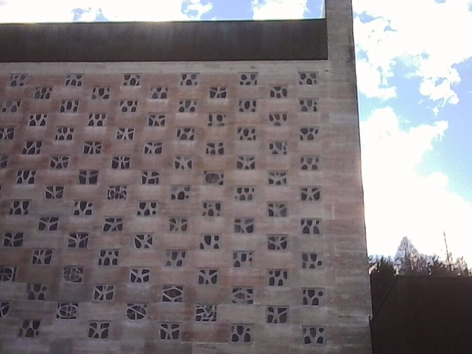 Concrete facade with a grid of small patterned openings, sunlight catching the upper edge against a pale winter sky.