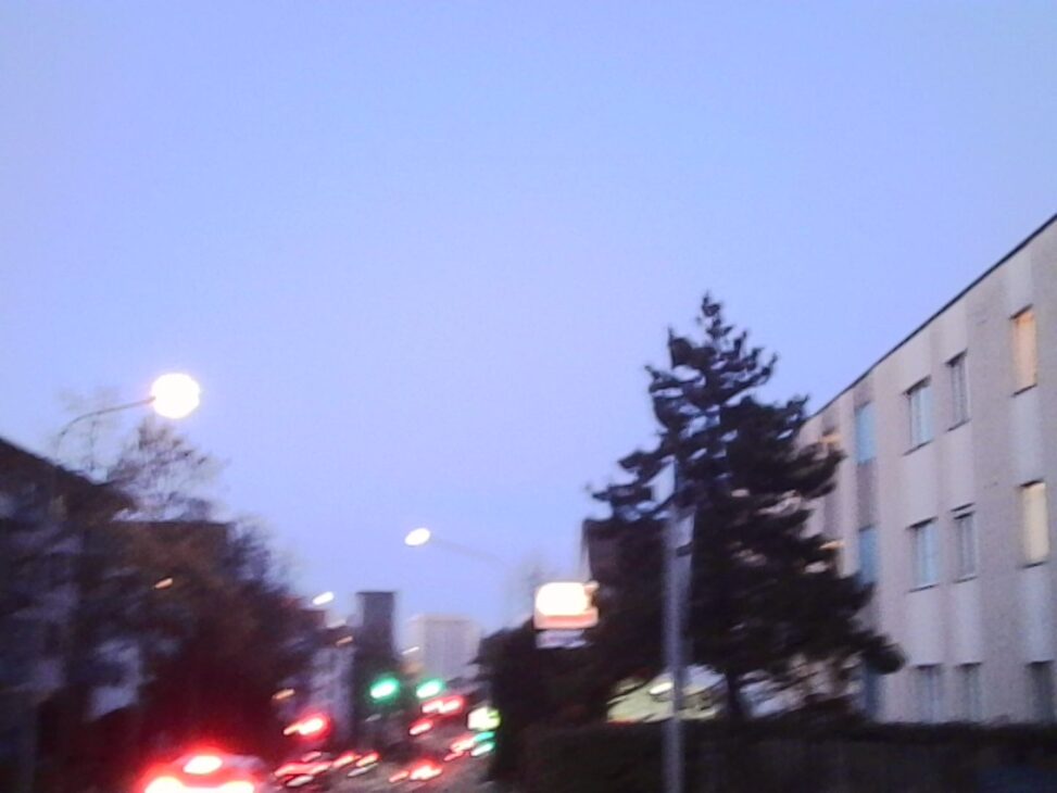 A low-angle, heavily blurred street scene at dusk. Glowing white streetlights and red taillights streak across a hazy blue sky. A dark evergreen tree and a pale apartment building frame the right side, capturing a sense of disorienting motion and physical fatigue.