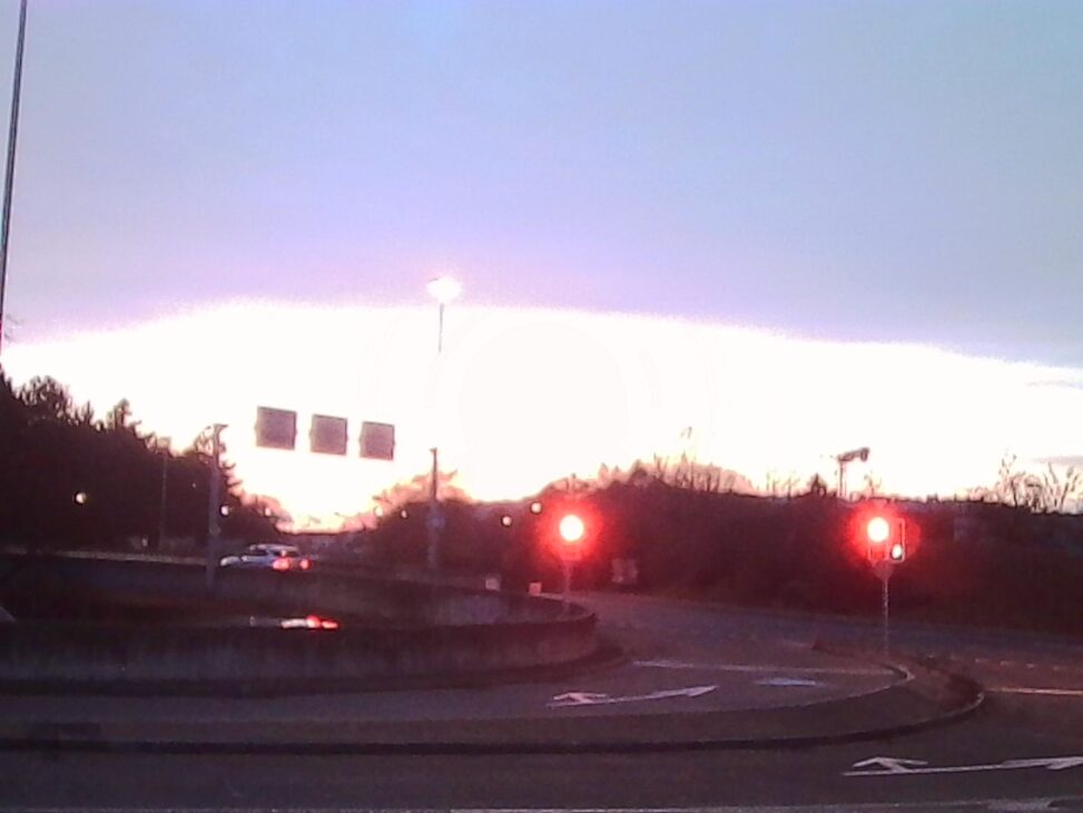 A wide, slightly soft-focus street scene at dusk near a road junction. Red traffic lights glow against a pale violet-blue sky, with dark tree lines and road markings silhouetted in the foreground. The scene conveys stillness, distance, and the quiet pause of evening.