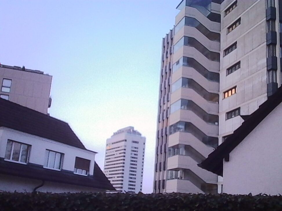 Low-angle view of Oerlikon high-rises at dusk. Older, lower residential buildings frame the foreground, while tall concrete apartment towers with zigzag balconies rise behind them against a pale blue evening sky, suggesting scale, fatigue, and quiet urban transition.