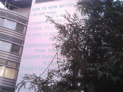 Facade of a building featuring Fischli/Weiss’ artwork “How to Work Better,” partially framed by tree branches in the foreground.