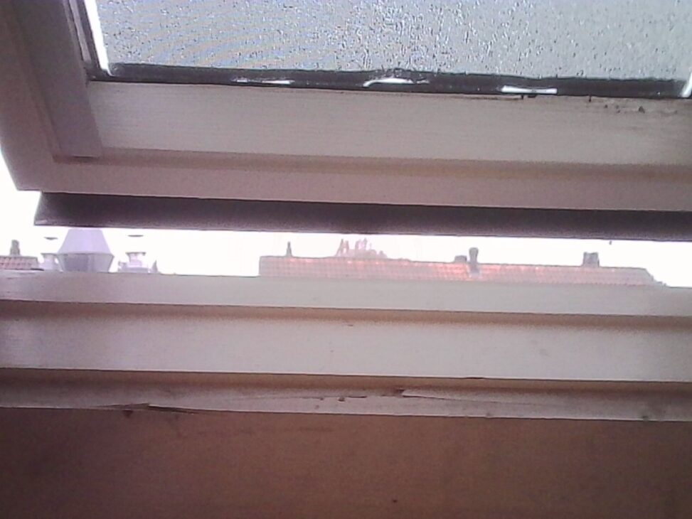 View through a partially open skylight window onto rooftops under an overcast sky, with condensation on the glass.