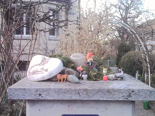 Small figurines arranged on a stone garden post, including animals and a seated character, with early spring foliage and residential buildings in the background.