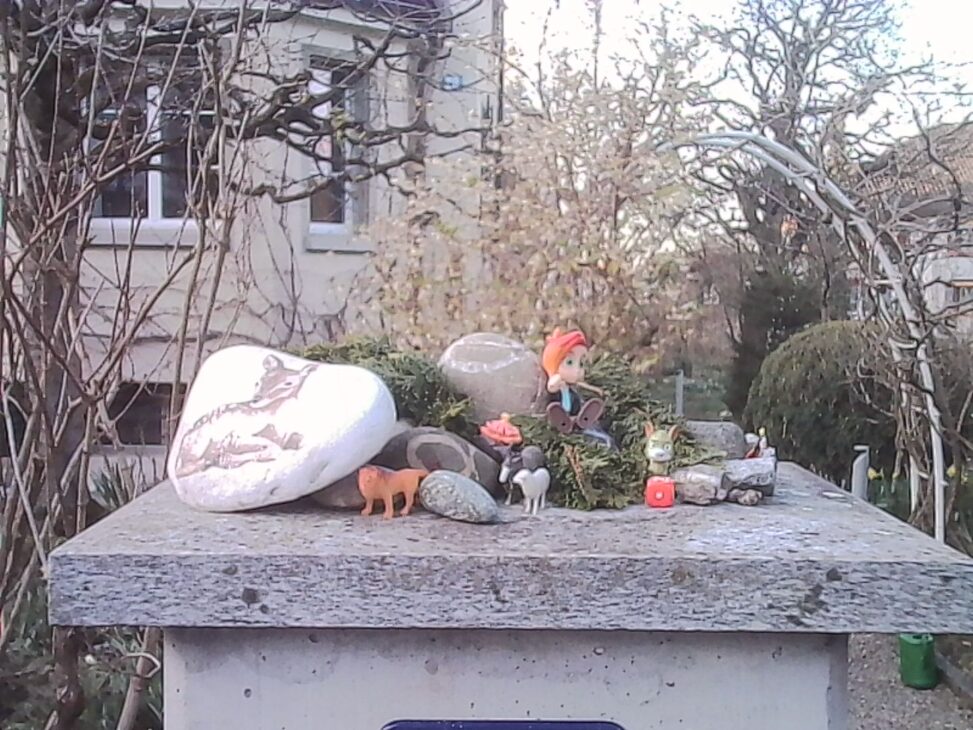 Small figurines arranged on a stone garden post, including animals and a seated character, with early spring foliage and residential buildings in the background.