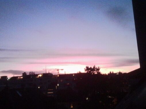 Dark silhouettes of rooftops and trees against a softly glowing pink and orange sunset on the western horizon, framed by the edge of a building on the right.