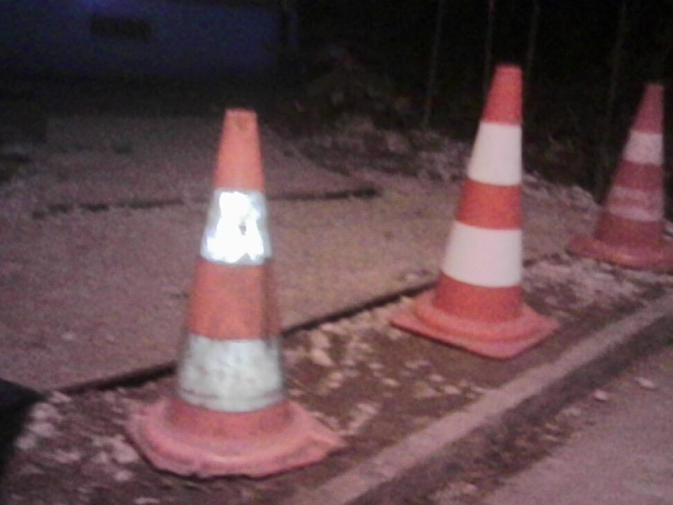 Three orange roadwork cones standing on a sidewalk at night, one with a reflective band glowing brightly under street lighting.