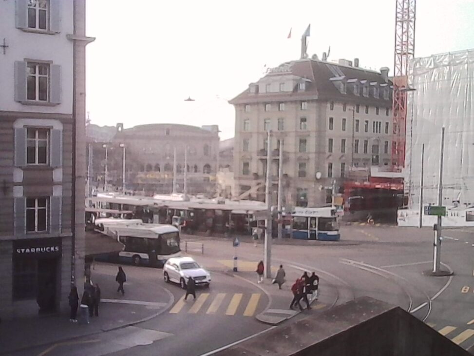 View over Zurich’s Central square with buses, pedestrians crossing a yellow-striped crosswalk, and historic buildings in bright afternoon light.