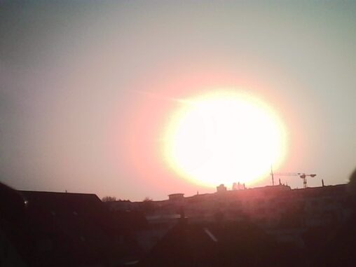 Bright setting sun over a Zurich skyline, with silhouettes of buildings and construction cranes against the glowing sky.
