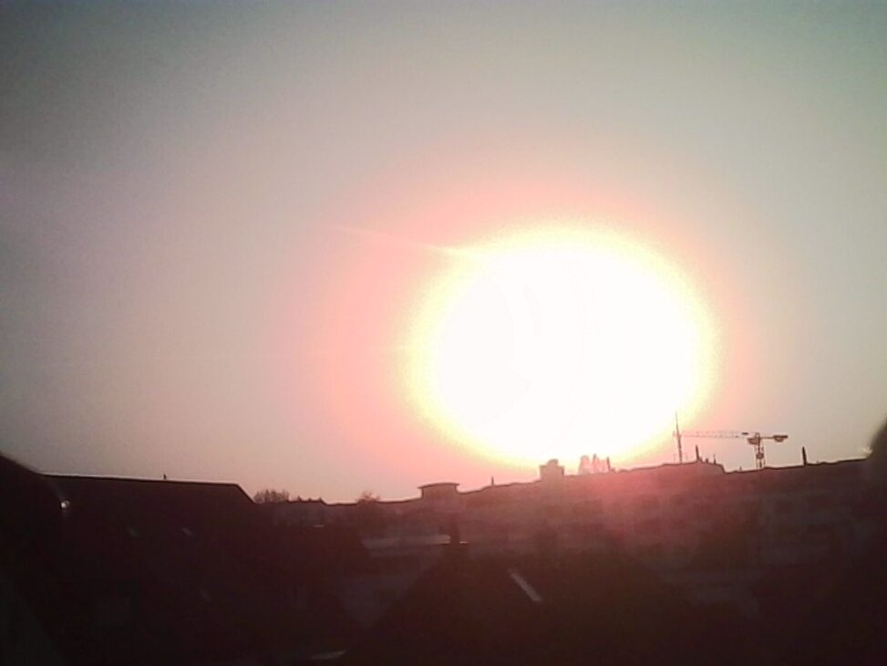 Bright setting sun over a Zurich skyline, with silhouettes of buildings and construction cranes against the glowing sky.