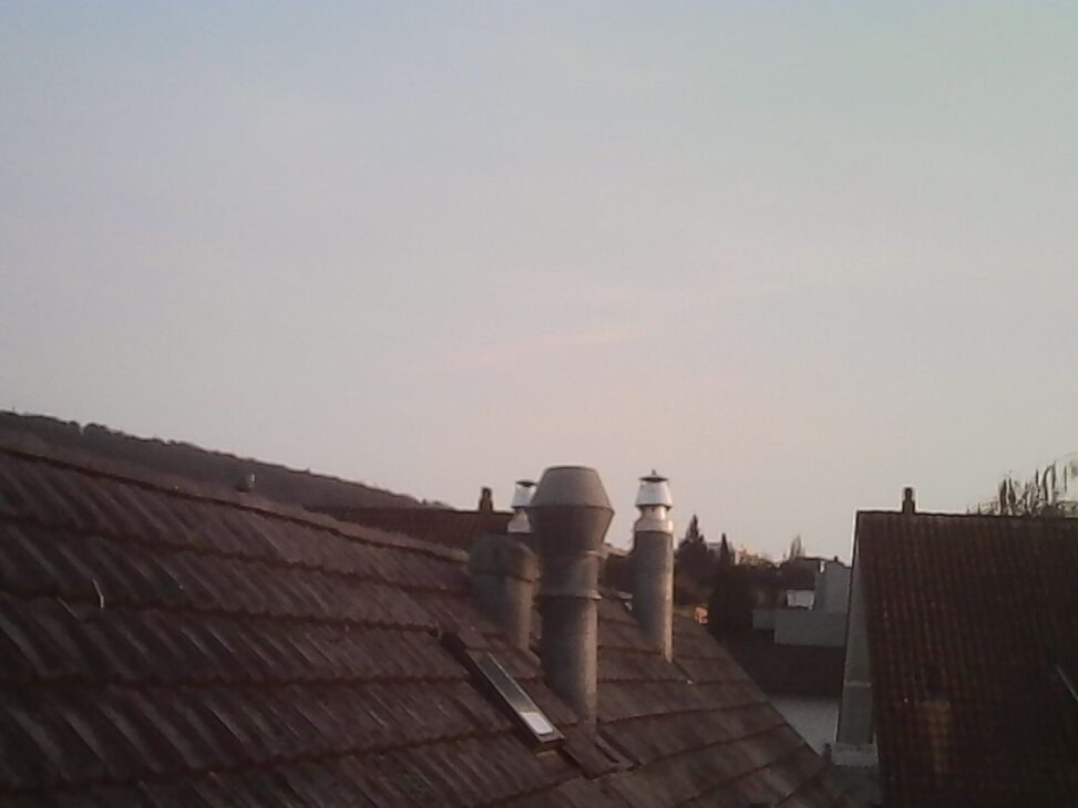 Rooftops with clay tiles and two chimney stacks shot from a window, pale blue sky above, soft warm light suggesting early spring