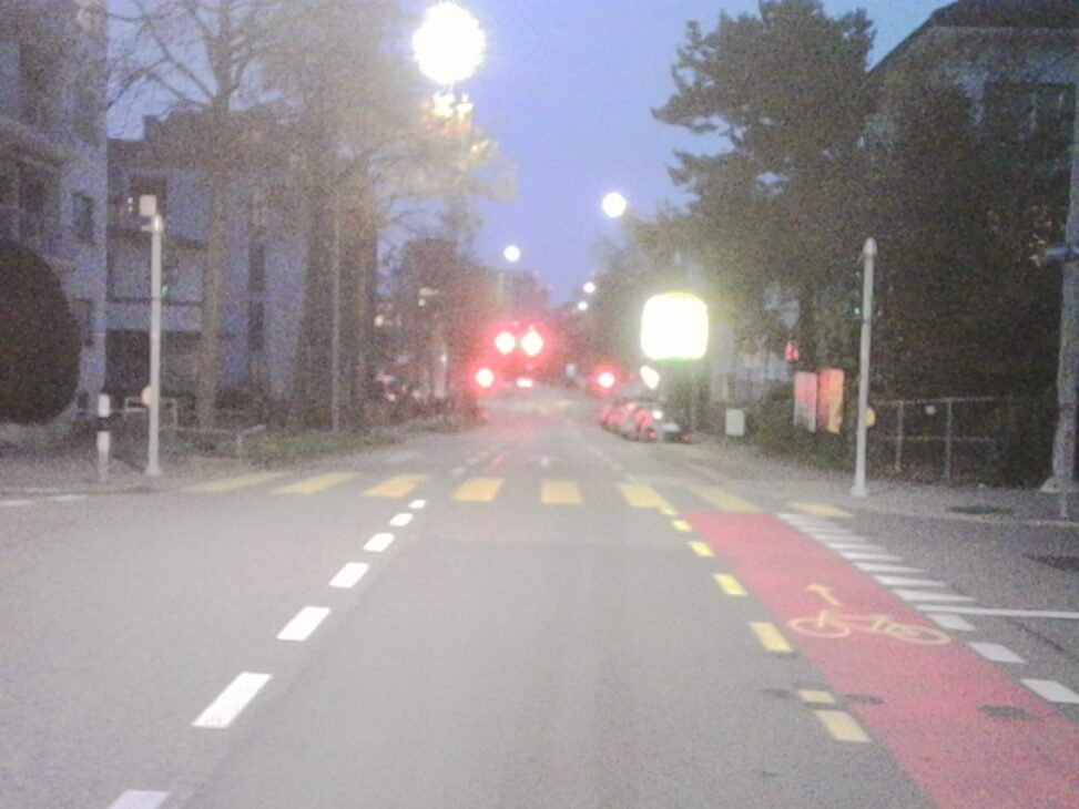 Dusk view down a quiet Zurich residential street toward Seebach, with glowing street lamps, a red bike lane, and traffic lights shining in the distance.