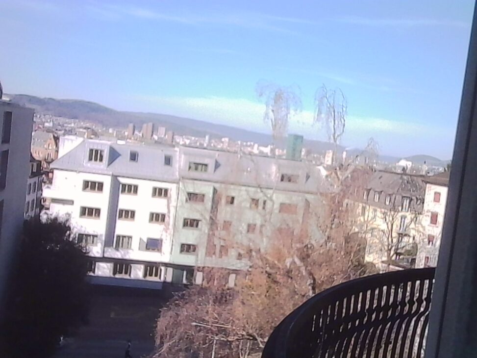 View across residential buildings toward the hills surrounding Zürich, photographed from a hospital window on a clear morning.