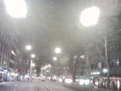 A wide city street at night lined with bright streetlights, shops, and buildings, with cars and pedestrians blurred by low light.