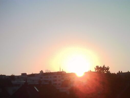 Sunset over a city skyline with the sun resting on the horizon, casting a bright halo and warm light over silhouetted buildings.