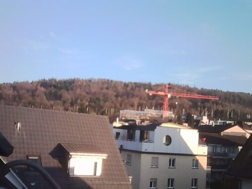 View over rooftops in Zurich toward Zurichberg hill under clear blue sky, with a construction crane and early spring light illuminating the scene.