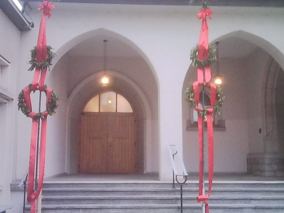 Arched church entrance framed by two tall Palm Sunday poles decorated with greenery and long red ribbons, with warm light glowing inside.