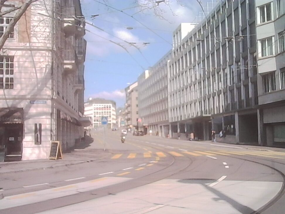 Curving tram tracks at Stampfenbachplatz in Zurich, with quiet streets, scattered pedestrians, and soft daylight breaking through clouds.