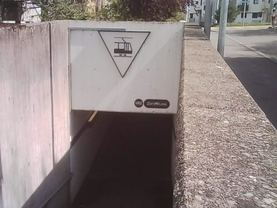 Triangular tram tunnel sign marking the underground “Tierspital” stop in Zurich, positioned above a dark underpass entrance along a concrete walkway in daylight.