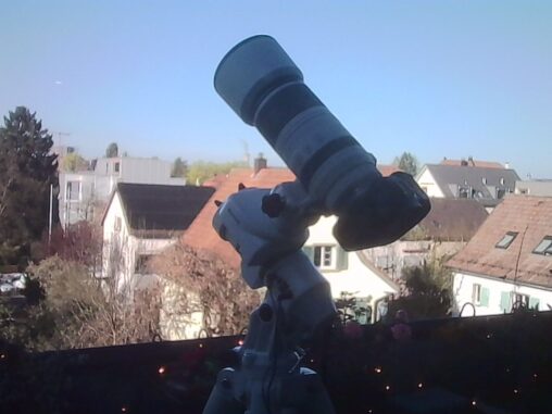 Camera with a large telephoto lens mounted on a star tracker on a balcony, with residential houses and a clear blue sky in the background