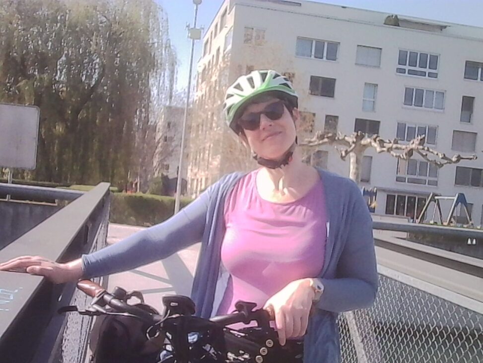 Smiling woman with a bicycle, wearing a helmet and sunglasses, leaning on a bridge railing in bright sunlight in Opfikon/Glattpark.