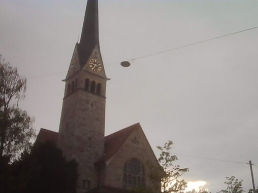 Church tower with a clock rising into a grey, overcast sky, partially framed by trees.