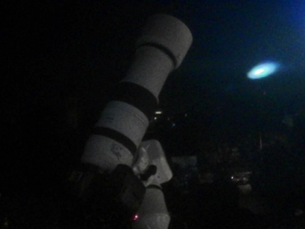 Low-light photo of a telescope setup outdoors at night, pointed upward, with faint lights in the background.