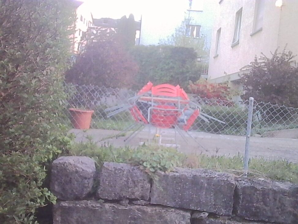A small pile of red garden chairs stacked and tangled behind a chain-link fence, seen from a slightly elevated angle. In the foreground, a low stone wall with patches of grass and weeds leads into a quiet residential garden scene, softly washed out by bright evening light.