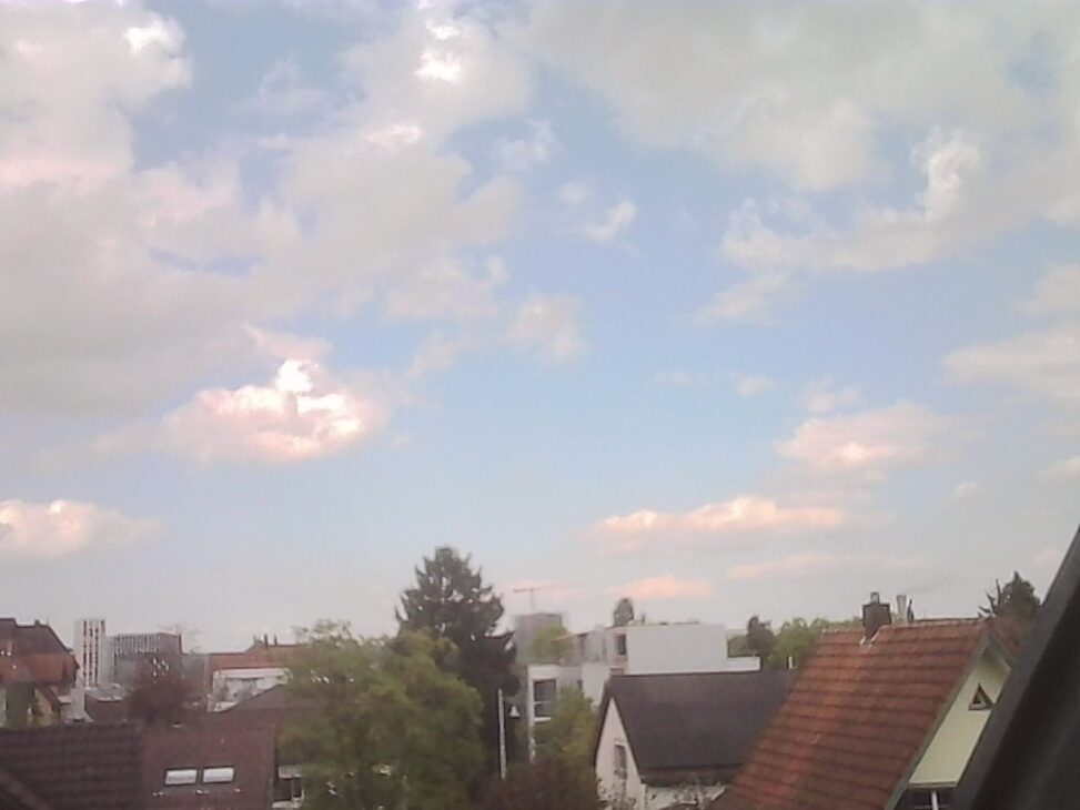 Soft, hazy sky with scattered clouds above a quiet residential neighborhood, rooftops in the foreground and the sun diffused behind thin cloud cover.
