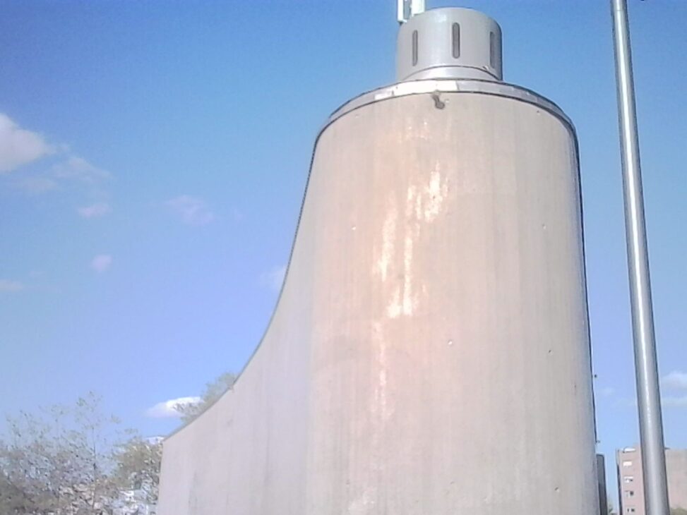 Concrete ventilation shaft rising from a curved highway cover, shaped like a smooth funnel against a pale blue sky.
