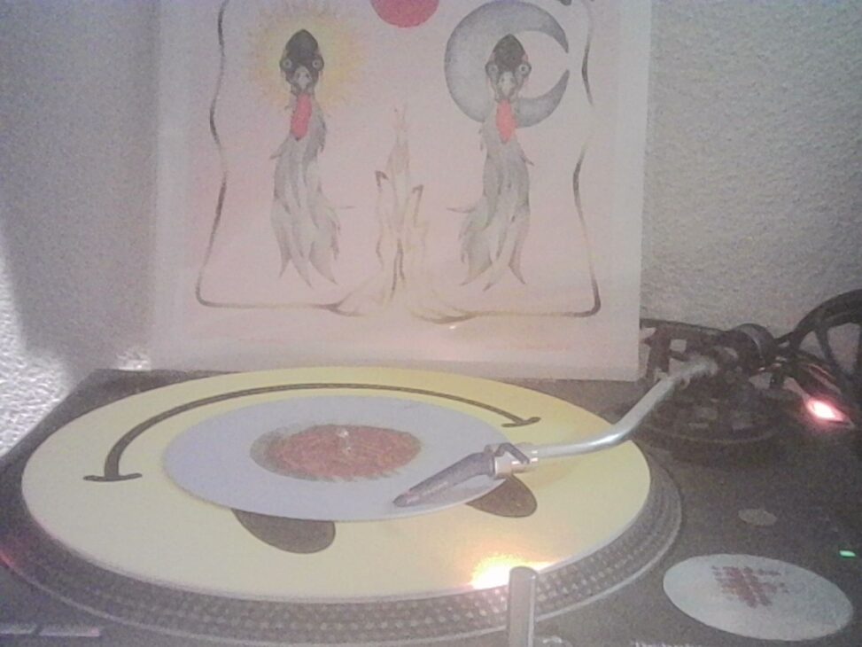 A turntable playing a vinyl record with a tonearm lowered onto the groove, illuminated by a small warm light, with a record sleeve featuring two illustrated figures in the background.
