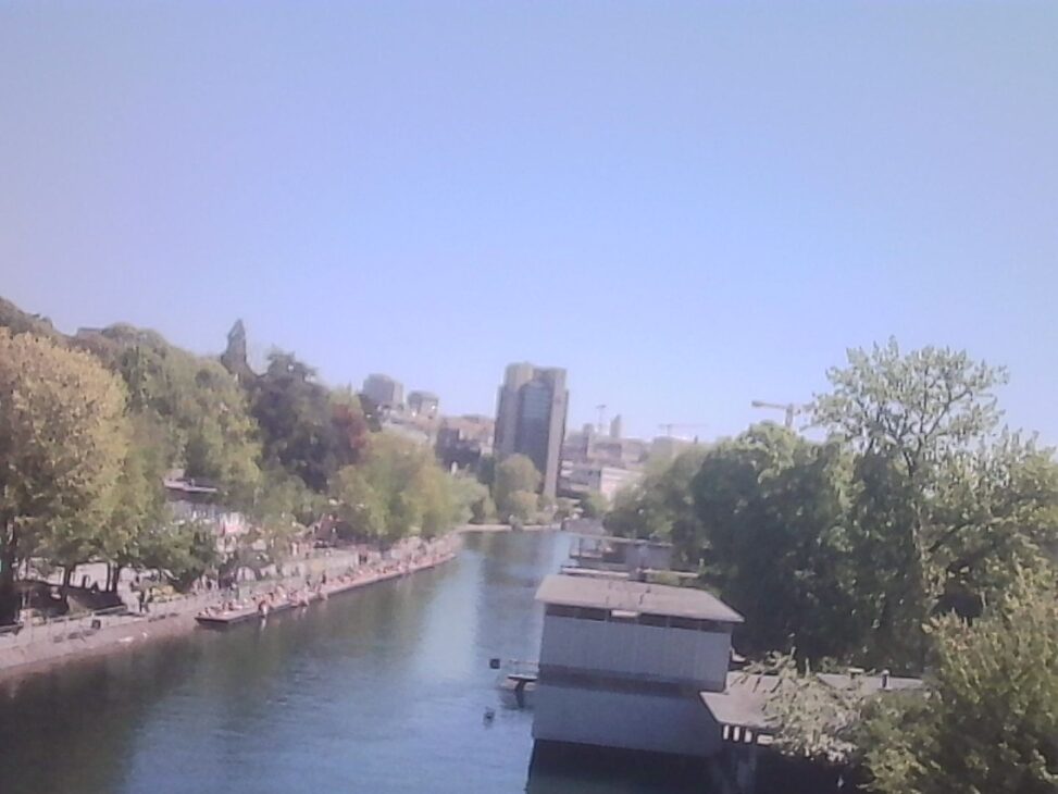 View from a bridge over the Limmat on a clear sunny day, with people lining the western riverbank, a boathouse in the foreground, and the city skyline and construction cranes visible in the haze.