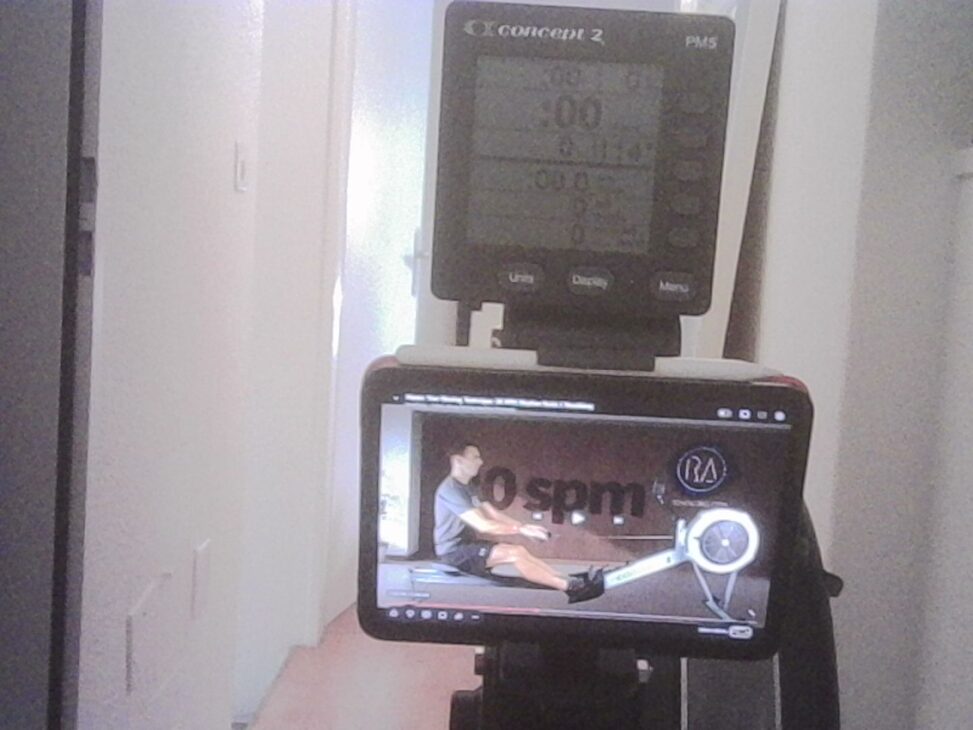 Concept2 rowing machine with a PM5 monitor and a tablet showing a rowing technique video, set up in a home environment.